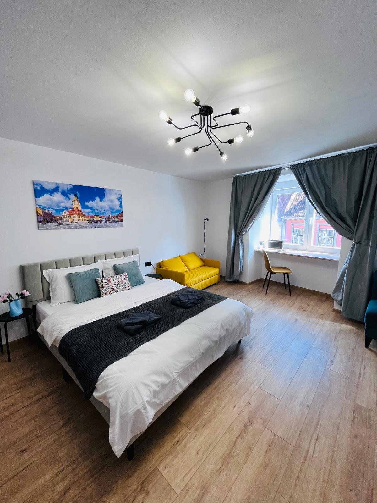 A cozy bedroom features a spacious bed dressed in white linens, complemented by decorative pillows. A modern chandelier illuminates the space, where a yellow armchair, a chair, and a large window with gray curtains add comfort and style.