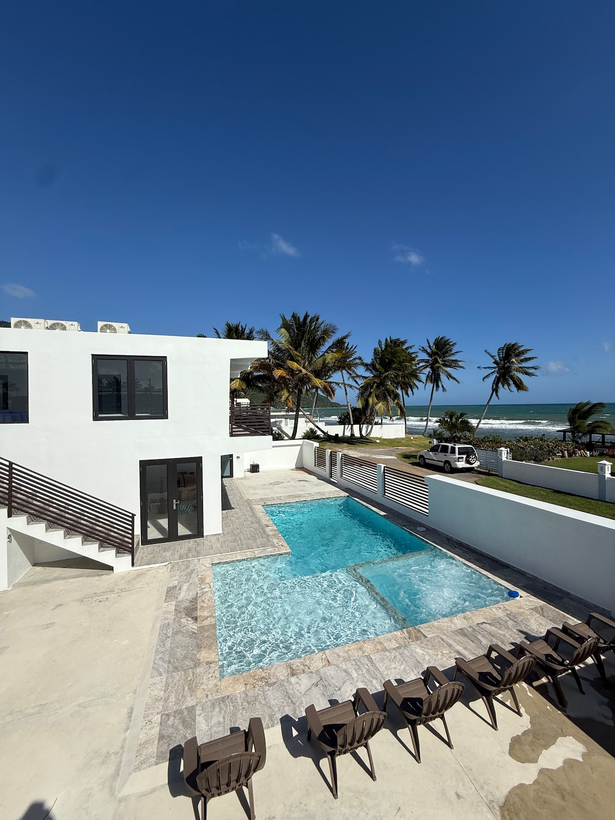 A spacious outdoor area features a heated pool with clear water, surrounded by a tiled patio. Comfortable lounge chairs line one side, while palm trees and ocean views complete the serene setting. The modern structure of the home is visible in the background, highlighting clean architectural lines.