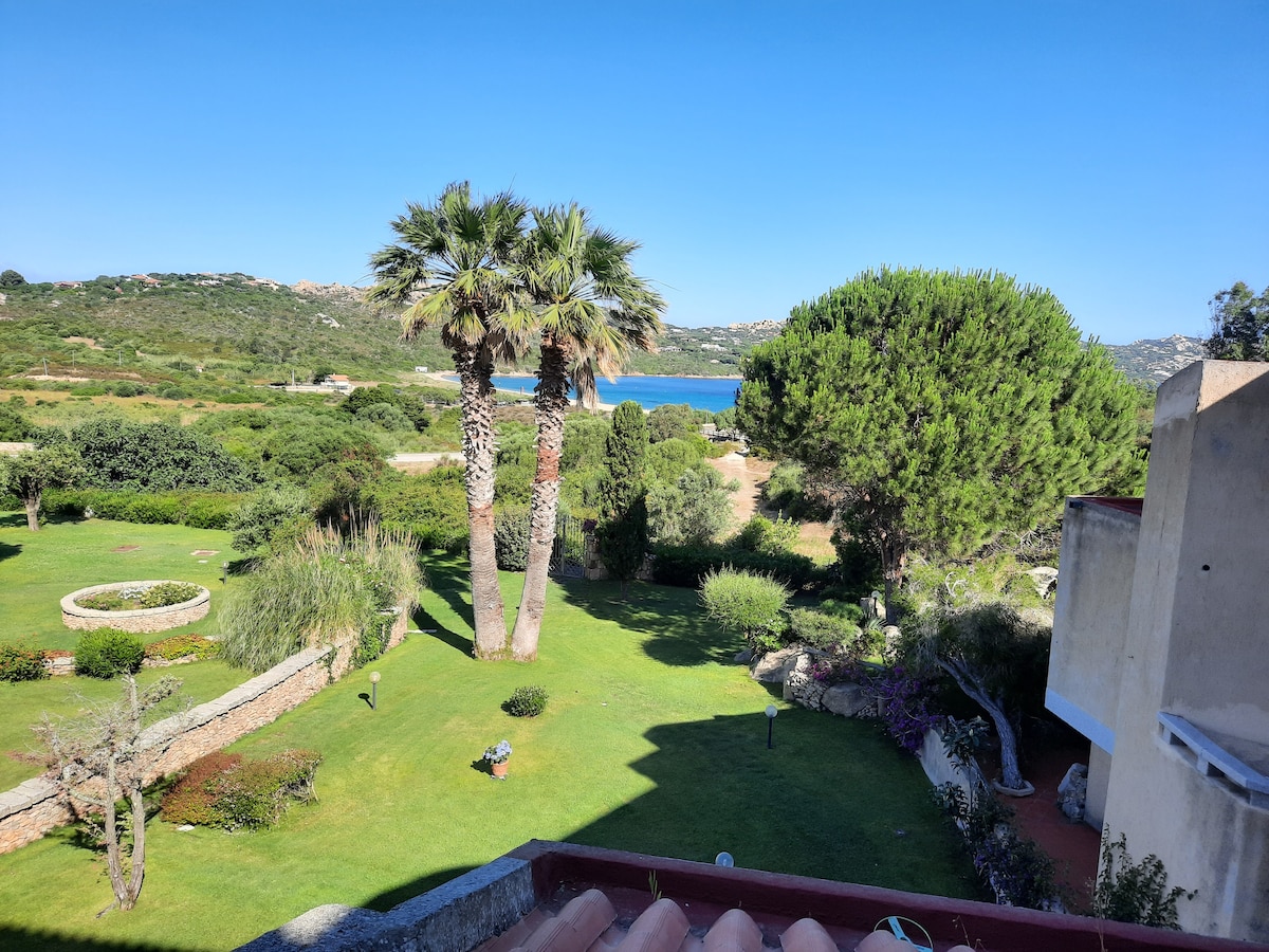 A lush garden area is visible, featuring palm trees and well-maintained greenery. Beyond the garden, a tranquil view of the water can be seen, framed by hills in the background under a clear blue sky.