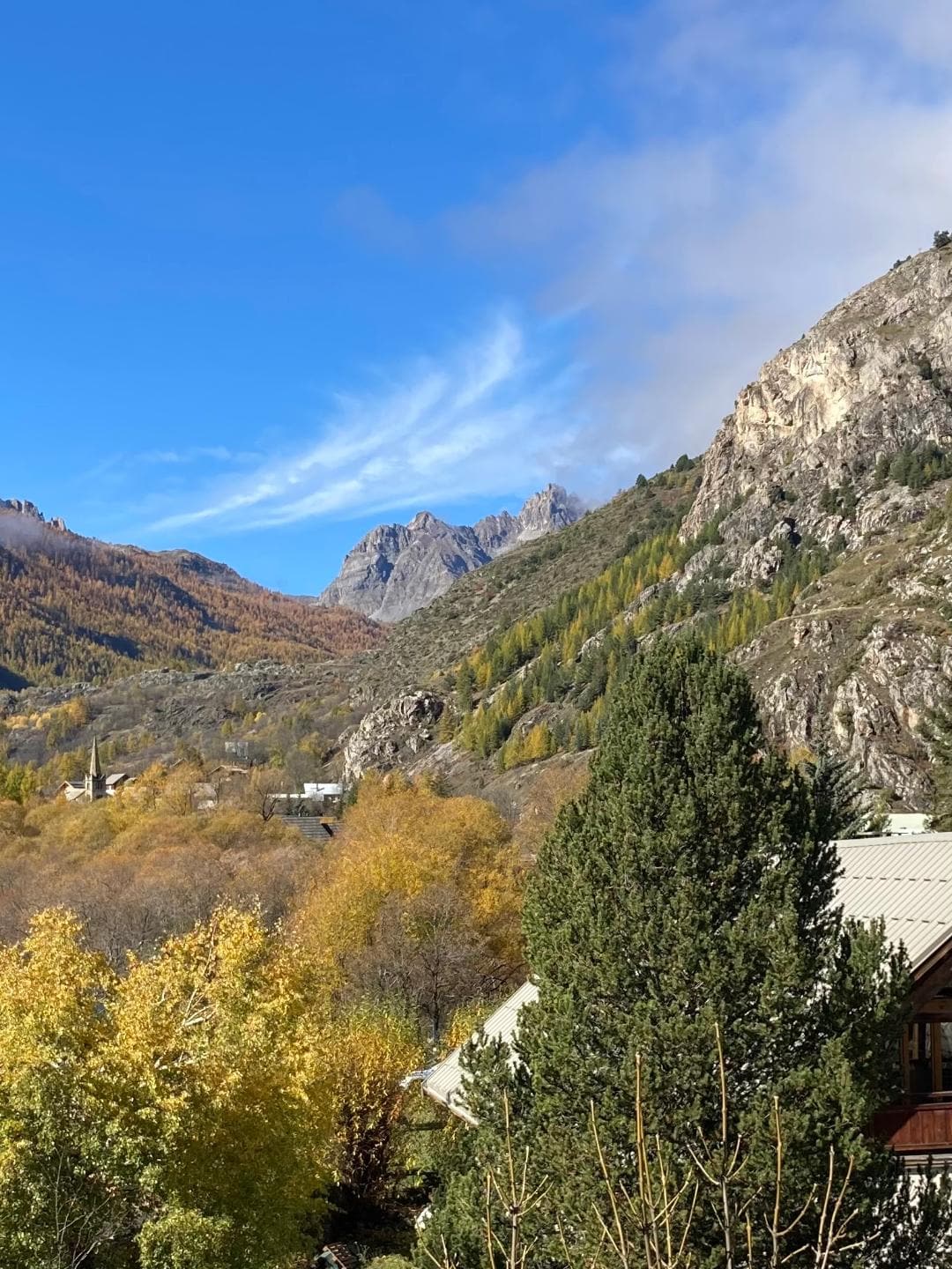 A scenic mountain view is presented, highlighting the surrounding peaks and valleys. The landscape features a mix of rocky terrain and trees displaying autumn colors, with a clear blue sky overhead. The peaceful environment conveys the natural beauty of the location.
