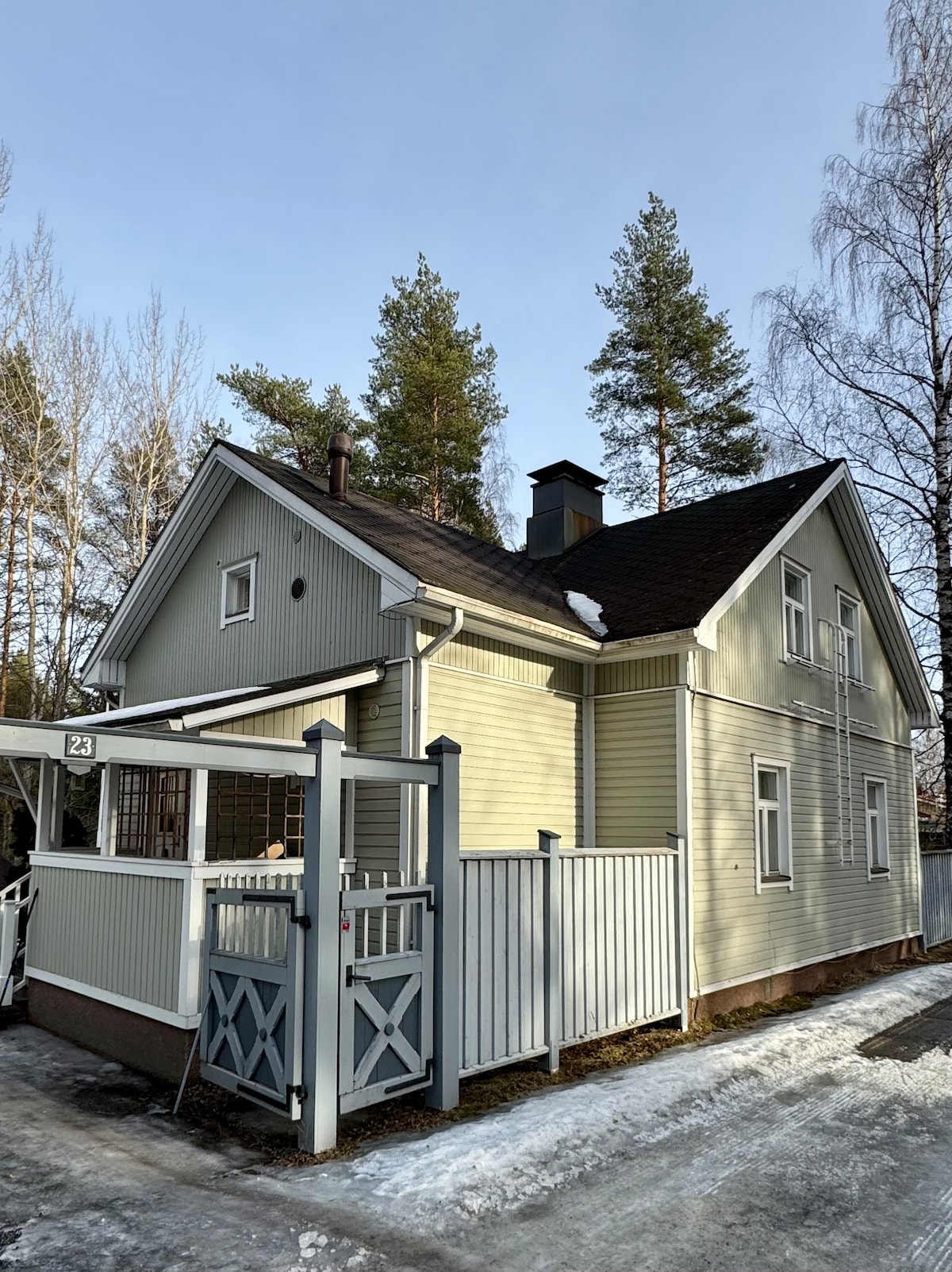 The exterior of a renovated wooden house is displayed, featuring light green siding and a dark roof. Tall pine trees surround the property, and a gated entrance is visible at the front. Bare patches of grass and remnants of snow can be seen on the ground.