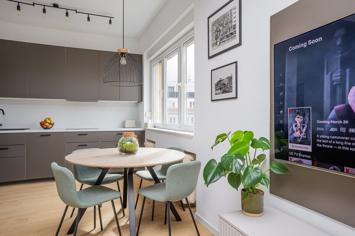 A modern kitchen area features sleek cabinetry and a round dining table surrounded by four chairs. A green plant adds a touch of nature, while the wall-mounted TV displays upcoming programs. Natural light flows in through large windows, creating a bright and welcoming space.
