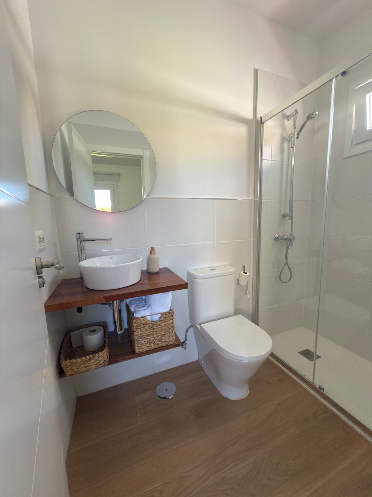 A bright bathroom features a round mirror above a modern sink with a wooden shelf underneath. A shower with glass doors is present, along with a toilet. Light wooden flooring complements the overall design, creating a clean and minimalistic environment.