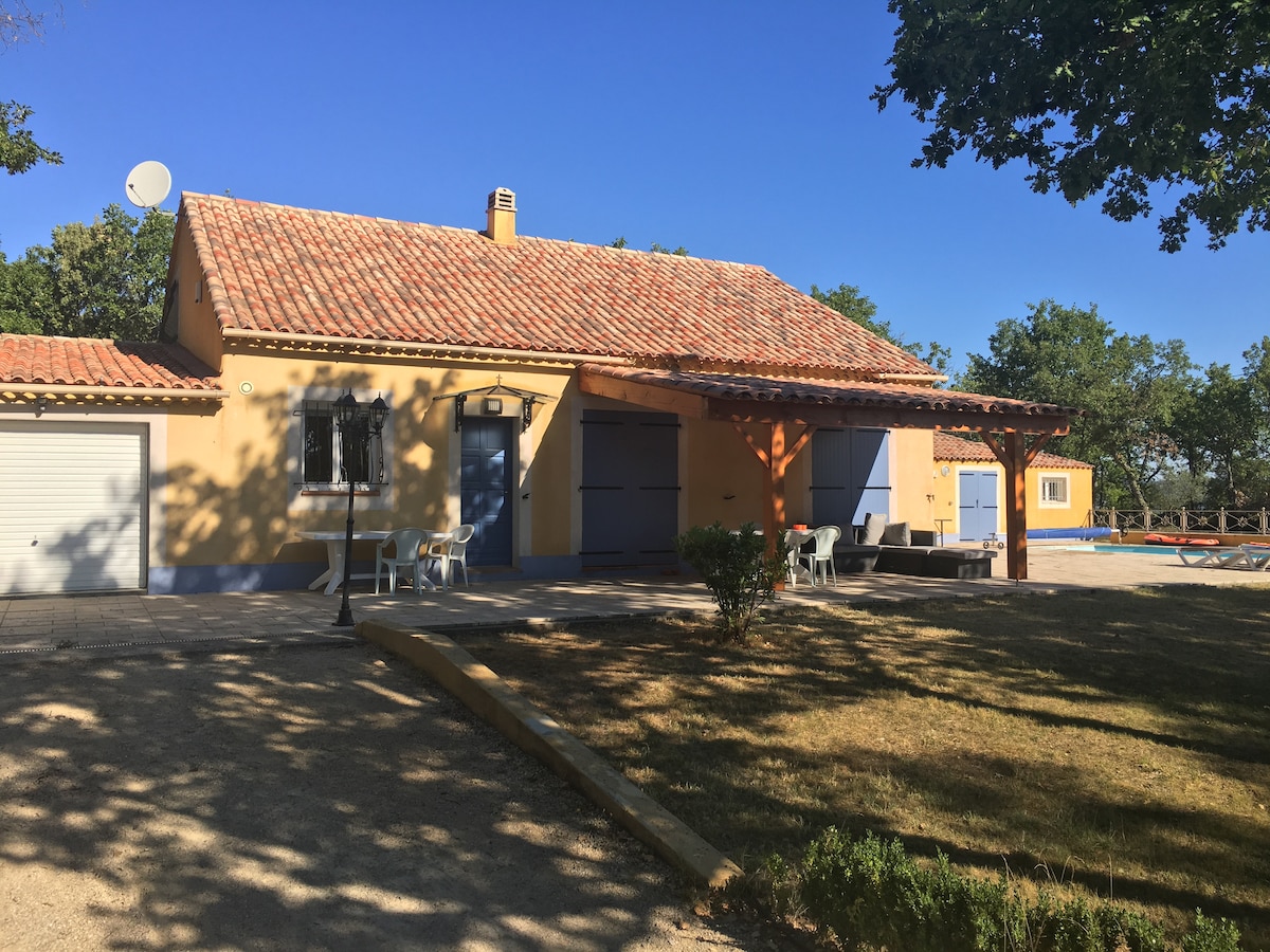 A single-story home is set on a grassy area, framed by trees. A covered terrace with seating is visible, offering shaded outdoor space. The house features a tiled roof and a garage, with a clear blue sky above enhancing the inviting exterior.