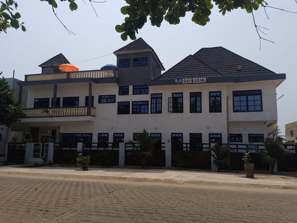 The exterior of a multi-story building is shown, featuring a modern design with multiple gabled roofs. Large windows allow natural light to fill the space. Lush greenery is present around the property, and a bright orange umbrella is visible atop the structure.
