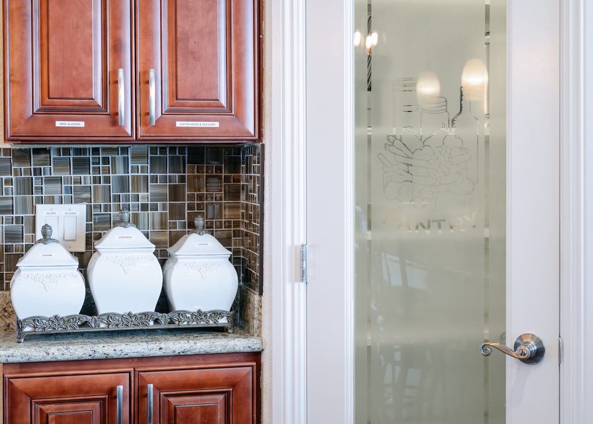 A well-appointed kitchen area features rich wooden cabinetry alongside a mosaic backsplash. Three decorative ceramic canisters are aligned on a polished granite countertop. A glass pantry door with etched design offers a glimpse into additional storage space.