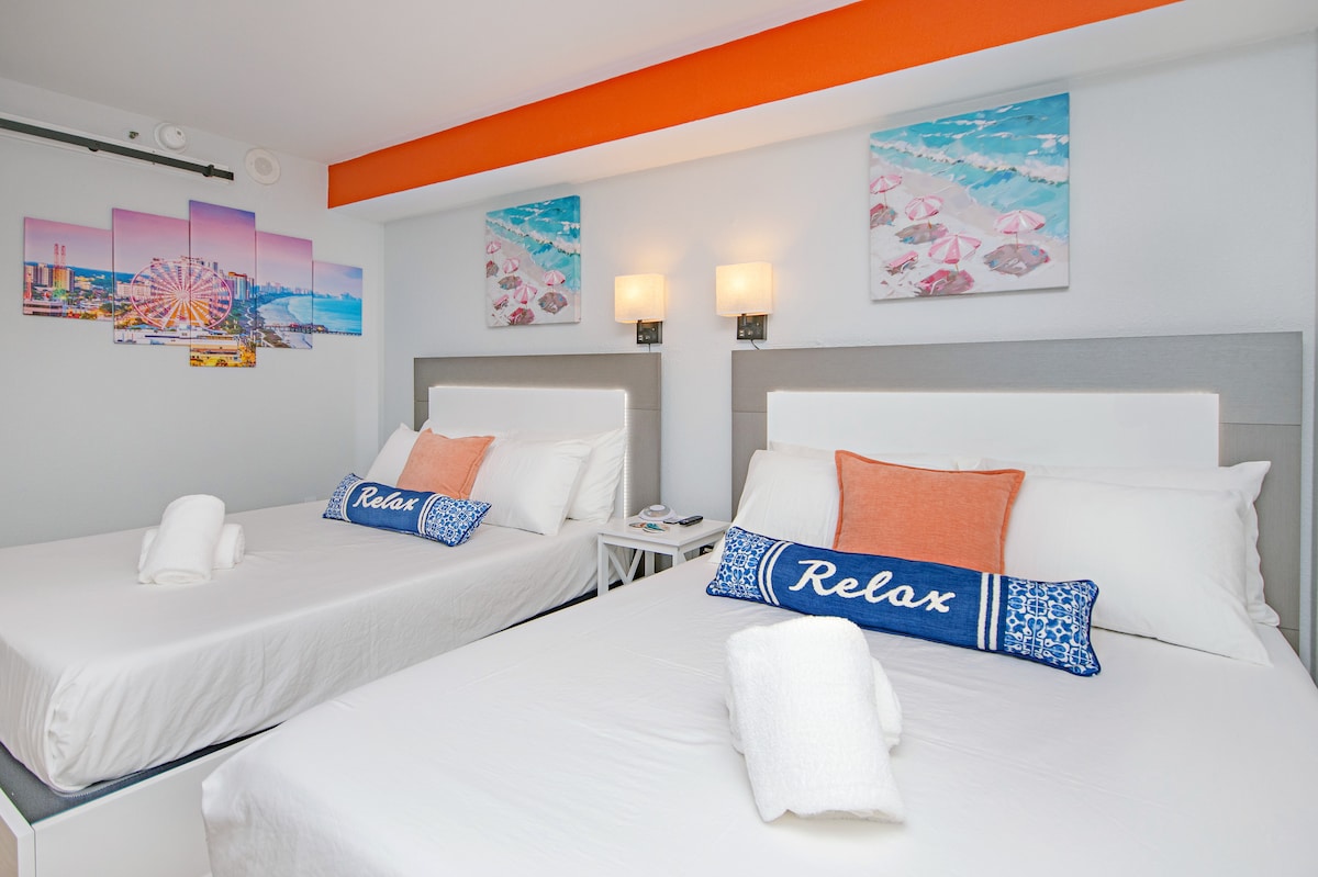 A spacious room features two queen beds with crisp white linens and decorative pillows. The walls are adorned with vibrant beach-themed artwork, and soft lighting is provided by wall sconces. A welcoming touch is added with plush towels neatly arranged on the beds.
