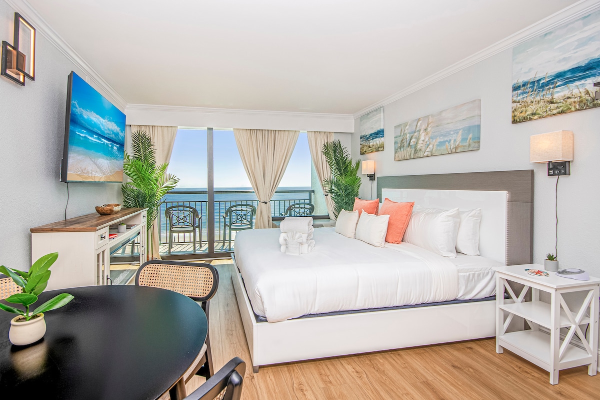A spacious bedroom features a king bed dressed in white linens with coral accent pillows. Light floods the room from large balcony doors offering ocean views. A cozy seating area, small dining table, and tropical decor complete this inviting coastal retreat.