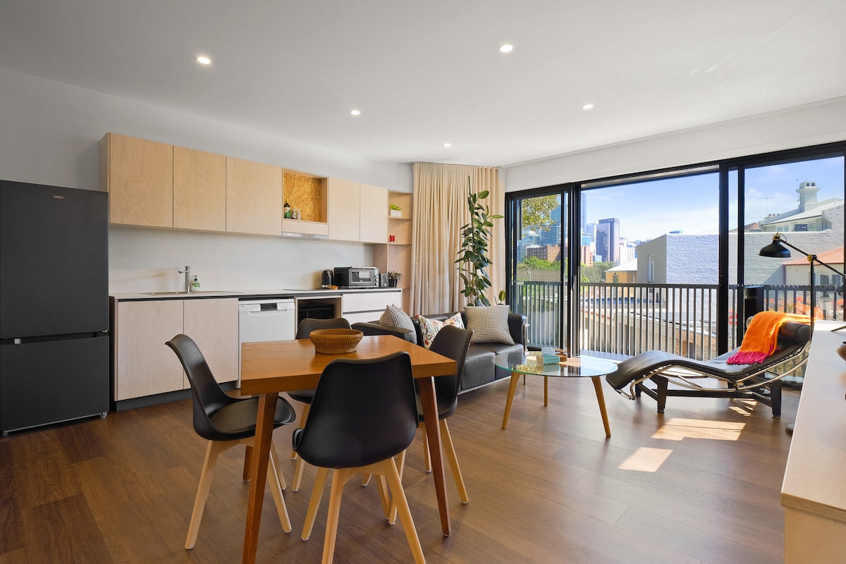 An open-plan living area features a wooden dining table surrounded by black chairs. A compact kitchen with light wood cabinetry and dark appliances is visible. A black leather sofa and a Le Corbusier lounge chair are positioned beside large windows, which provide views of the outdoors and natural light.