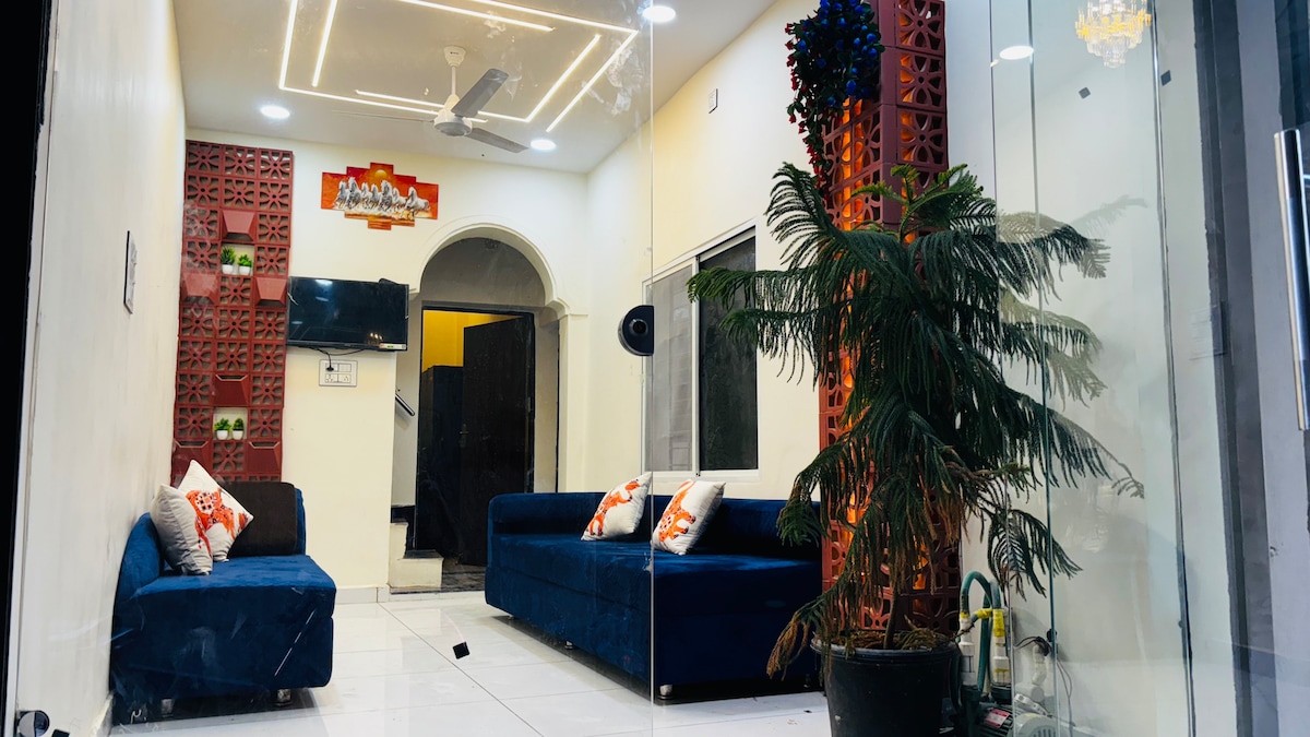 A modern living area features two blue sofas, surrounded by glass walls that create an airy setting. Decorative wall elements add texture, while a small indoor plant provides a touch of greenery. Soft lighting enhances the ambiance.