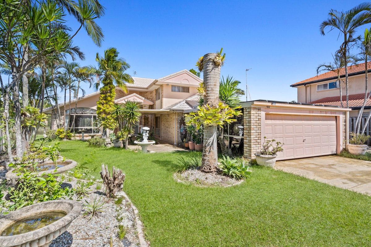 A well-maintained exterior showcases a charming two-story home surrounded by lush greenery. Palm trees and various plants frame the entrance, while a manicured lawn adds to the inviting appeal. A single-car garage is visible on the side, and a pathway leads through the vibrant garden.