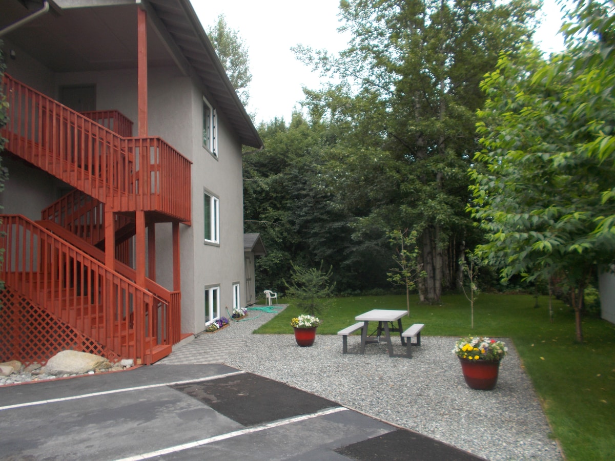 The exterior of a multi-story building is shown, featuring a red staircase leading to higher levels. A gravel area is depicted, with a picnic table surrounded by planters filled with flowers. Lush green trees create a serene backdrop, contributing to the inviting outdoor space.