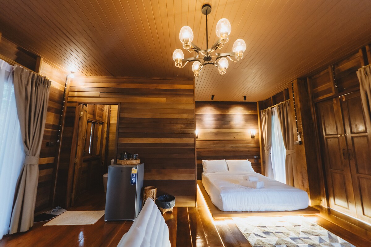 The interior features wooden walls and a light wood ceiling, creating a serene atmosphere. A spacious bed is positioned opposite a decoratively laid area rug, while a refrigerator and seating options are near the entrance. Natural light gently enters through large windows, enhancing the warm ambiance.