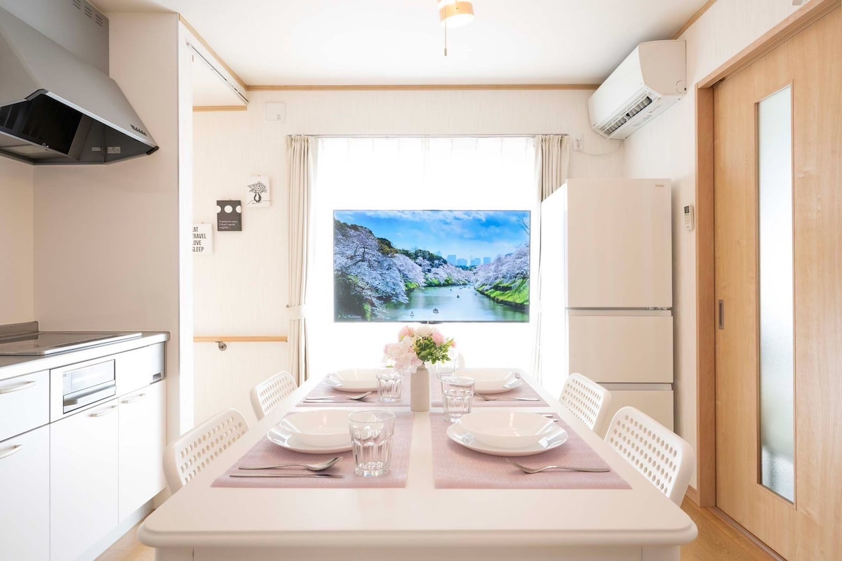 A dining area is featured with a rectangular table set for four, accompanied by white chairs. A large window allows natural light to fill the space, offering a view of a serene landscape. A refrigerator stands against the wall, next to a modern kitchen setup.