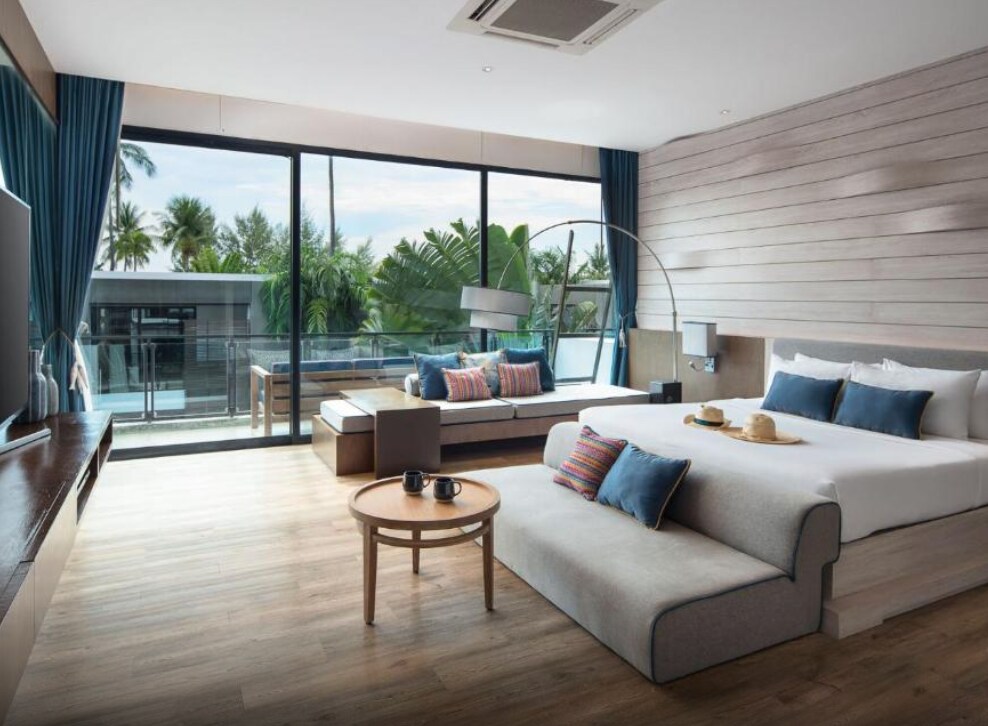 A spacious villa is showcased, featuring large glass doors that open to a private balcony. The room includes a cozy bed, a comfortable seating area, and a round table with two cups. Tropical plants and an outdoor setting are visible through the windows.