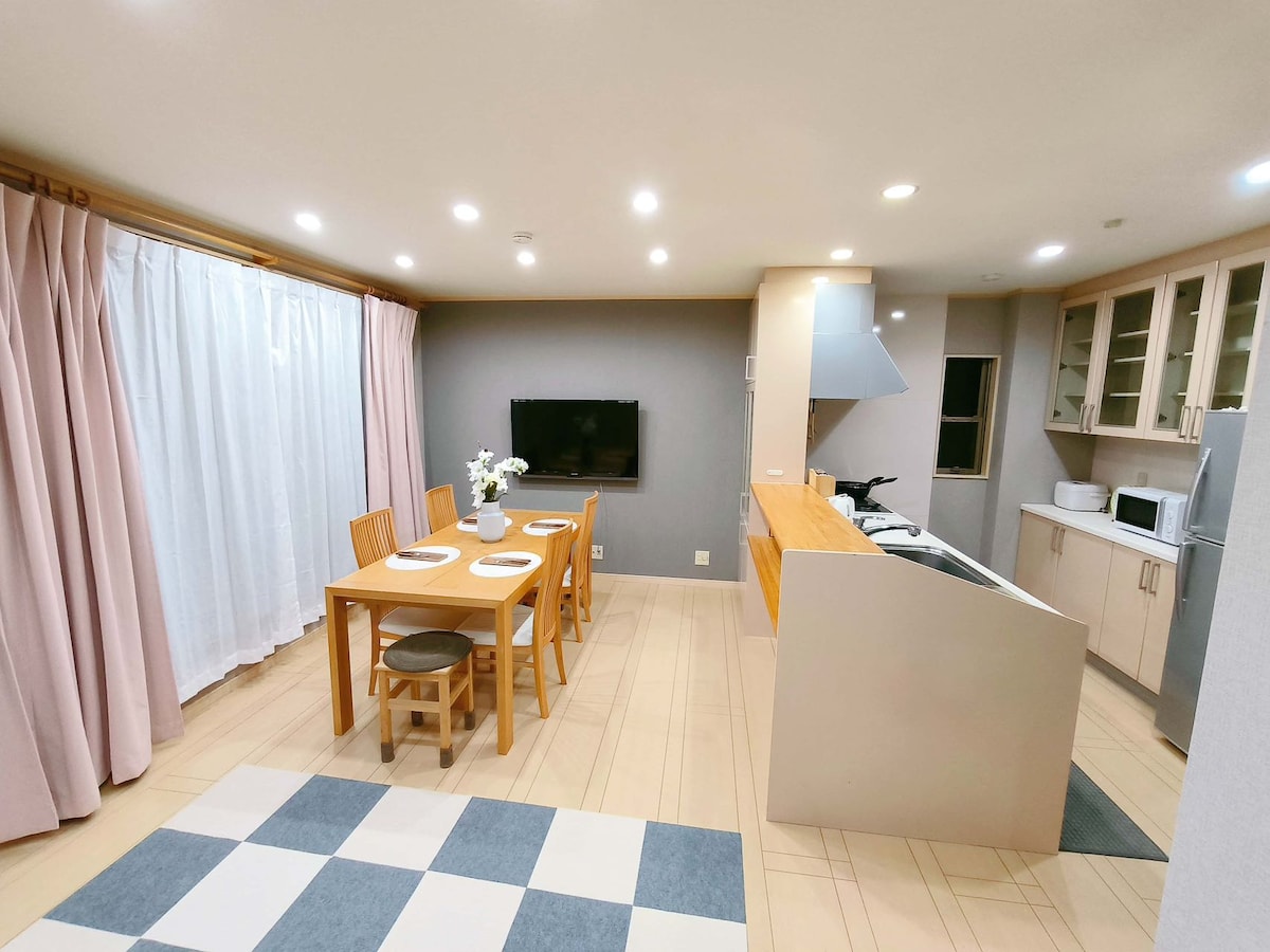 The open kitchen and dining area is characterized by a wooden dining table with four chairs, a television mounted on the wall, and a modern kitchen counter. Natural light filters through sheer white curtains, enhancing the spacious ambiance.