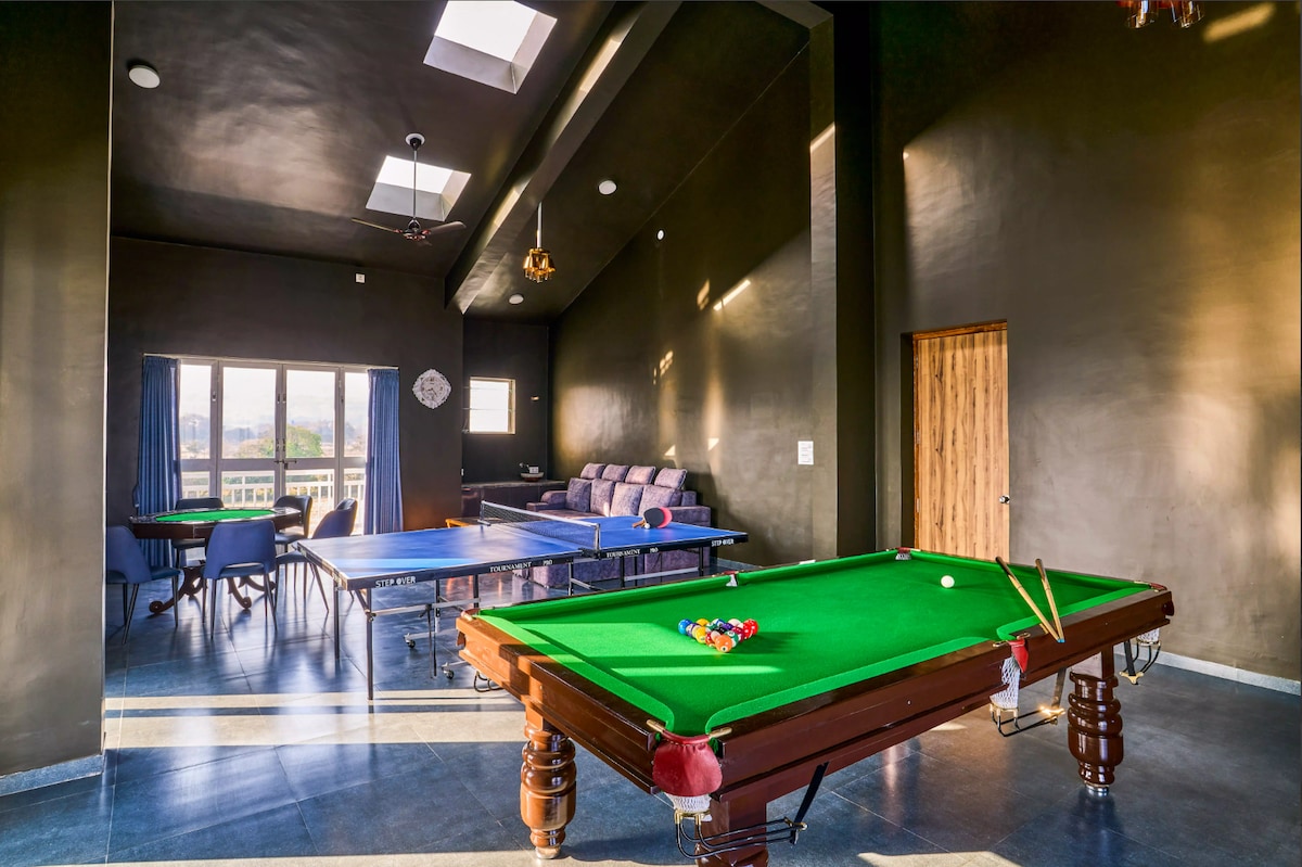 A games room featuring a billiard table with a set of colorful balls laid out. Natural light enters through skylights and a window, illuminating the dark walls and floor. A seating area with a sofa and dining table is visible in the background.