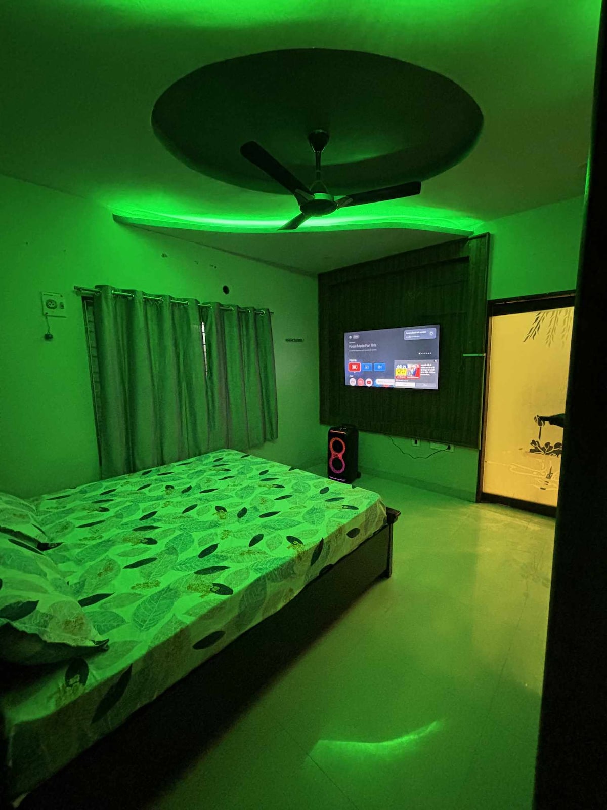 A bedroom features soft green lighting that creates a calm environment. A large bed is adorned with a patterned duvet cover. A wall-mounted television is visible, and a partially frosted glass door leads to another space. Curtains frame a window that allows natural light.