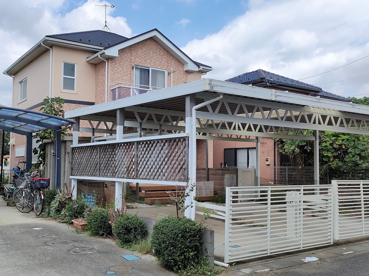 A two-story residence is featured, constructed with a combination of light-colored bricks and beige walls. A covered parking area is visible, providing space for vehicles. Lush greenery surrounds the exterior, and a wooden fence adds a touch of privacy. The sky is partly cloudy.