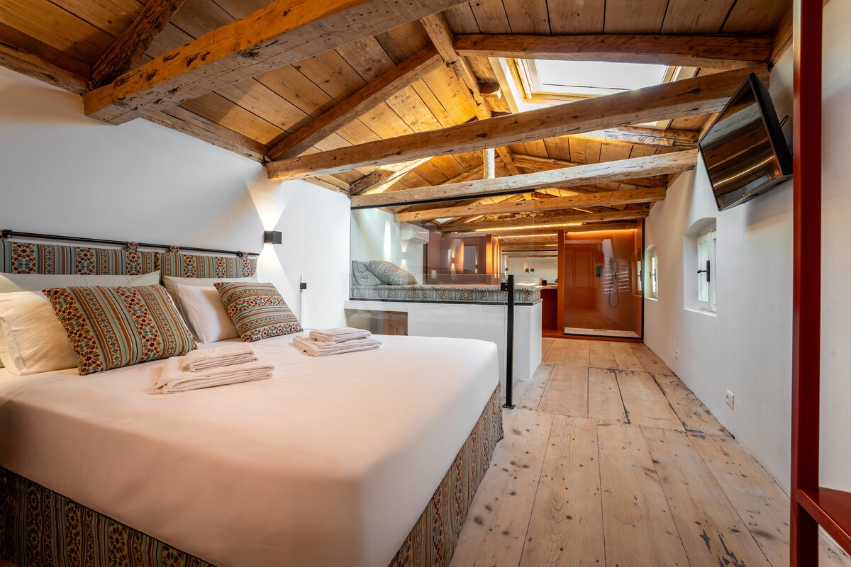 A spacious room features a large bed with patterned throw pillows, complemented by wooden beams overhead. Natural light floods the space through a skylight, illuminating the polished wood floor. A separate relaxation area is visible in the background, enhancing the room's openness.