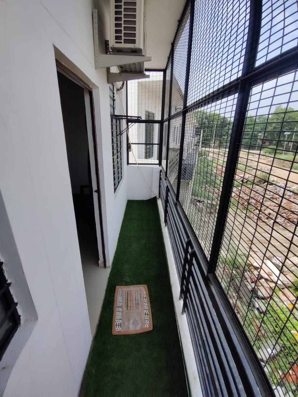 A narrow balcony is shown, featuring artificial grass covering the floor. One side displays a large window with metal grilles, allowing ample natural light. The balcony is accessible through an open doorway, providing a space for fresh air and outdoor views.