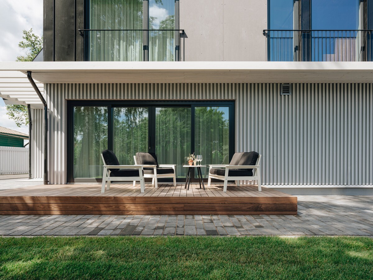 An outdoor terrace features comfortable seating with black cushions and a small table. Large glass doors offer views into the interior, while a wooden deck extends over the well-maintained lawn. Sunlight illuminates the area, enhancing the inviting and open atmosphere.