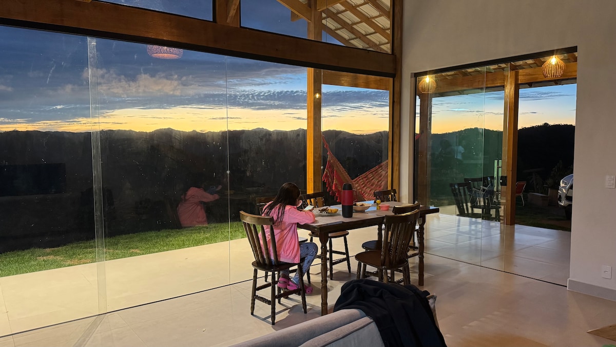 A spacious dining area features a wooden table surrounded by chairs, with a large glass wall providing a view of the sunset over the mountains. A hammock can be seen outside, enhancing the connection to nature.
