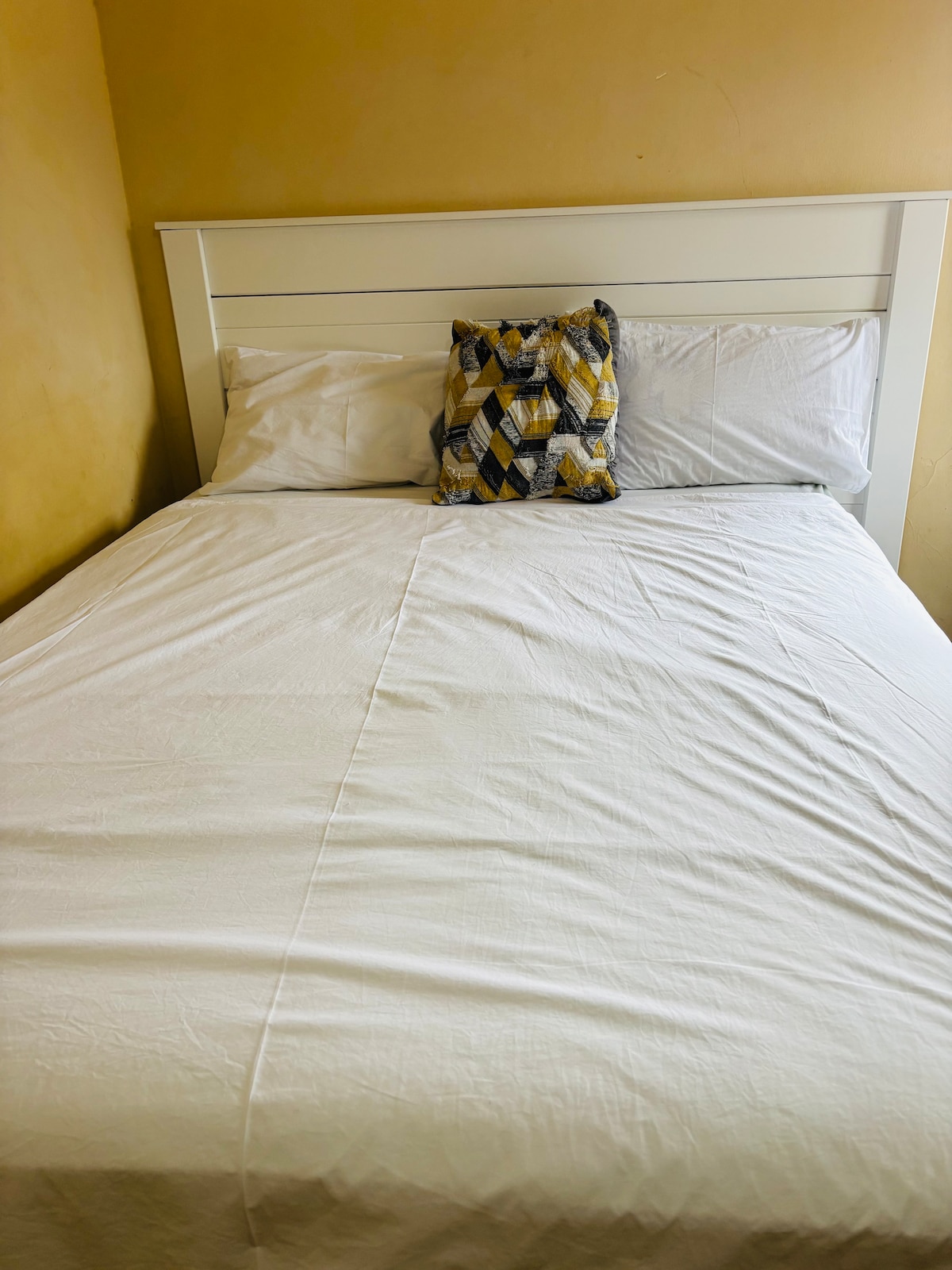 A spacious bed is presented with crisp white linens, accented by a decorative geometric pillow in yellow and navy. The walls exhibit a subtle warm tone, contributing to a calming atmosphere.