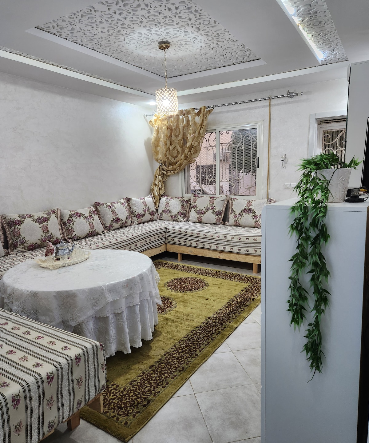 A cozy sitting area is furnished with a large, L-shaped couch covered in patterned fabric. A round table is centered, topped with a decorative cloth. Natural light filters through a window, while a green area rug adds warmth to the tile floor.