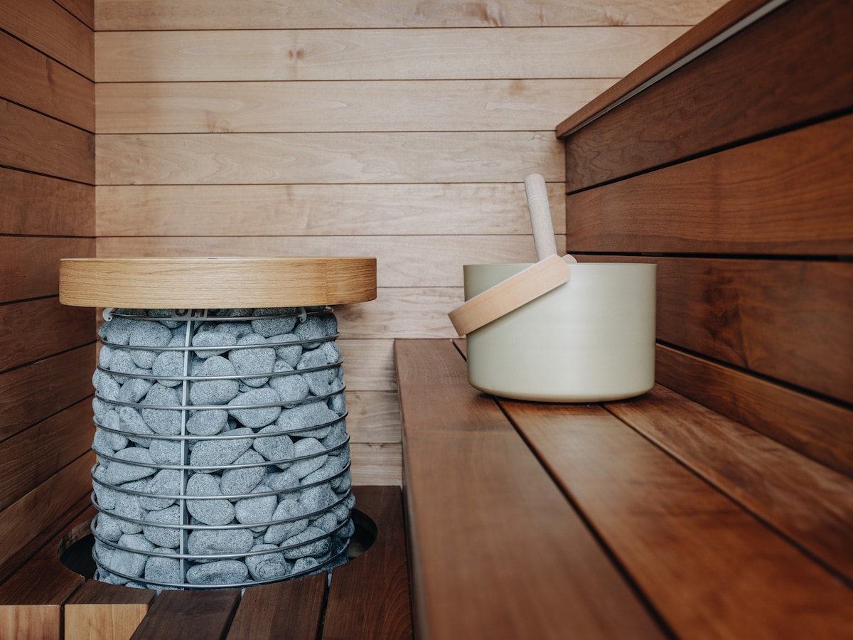 The interior of the sauna features warm wooden paneling and smooth wooden benches. A stone heater is positioned at the center, topped with a wooden seat. Adjacent, a simple ceramic bowl and wooden ladle are placed atop a wooden shelf, providing essentials for a soothing experience.