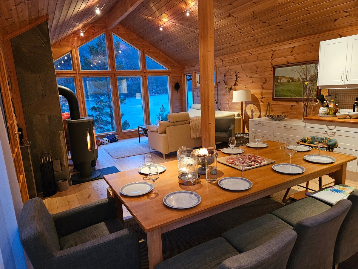 An open dining area is illuminated by natural light streaming through large panoramic windows. A spacious wooden dining table is set for a meal, surrounded by comfortable gray chairs. In the corner, a small wood stove adds warmth to the inviting space.
