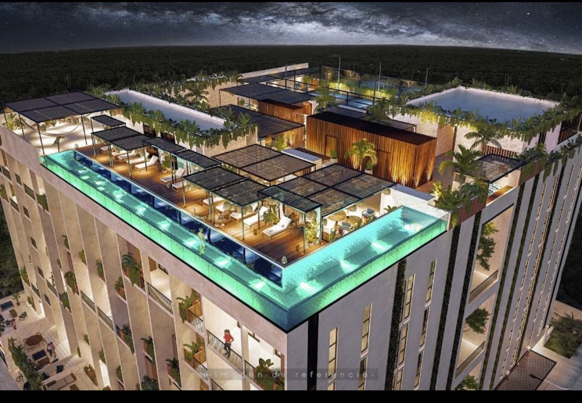 Aerial view of a modern building featuring multiple levels of terraces, surrounded by greenery. Rooftop amenities include an infinity pool, lounging areas with sunbeds, and shaded sitting spaces, offering a blend of relaxation and outdoor enjoyment.