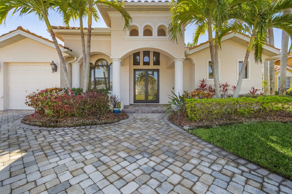 The homeās entrance is framed by tall palm trees and vibrant landscaping. A welcoming front porch features elegant columns and decorative arches. A neatly paved pathway leads to the double front doors, surrounded by colorful plantings and manicured grass.