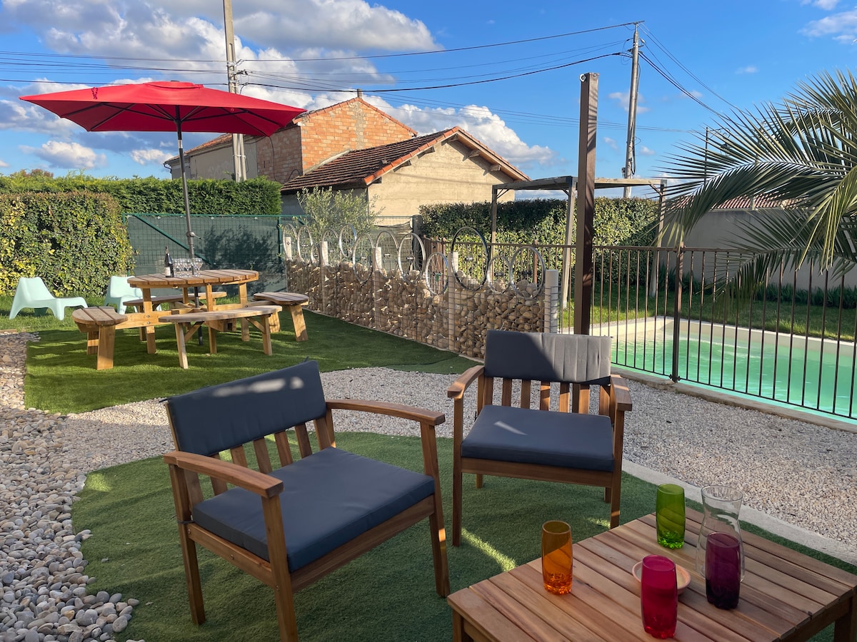 An outdoor area features two wooden chairs with cushioned seating, positioned on a grassy surface. A vibrant umbrella provides shade over a nearby picnic table. Beyond, a private pool is surrounded by a decorative stone wall and lush greenery, with a clear blue sky overhead.