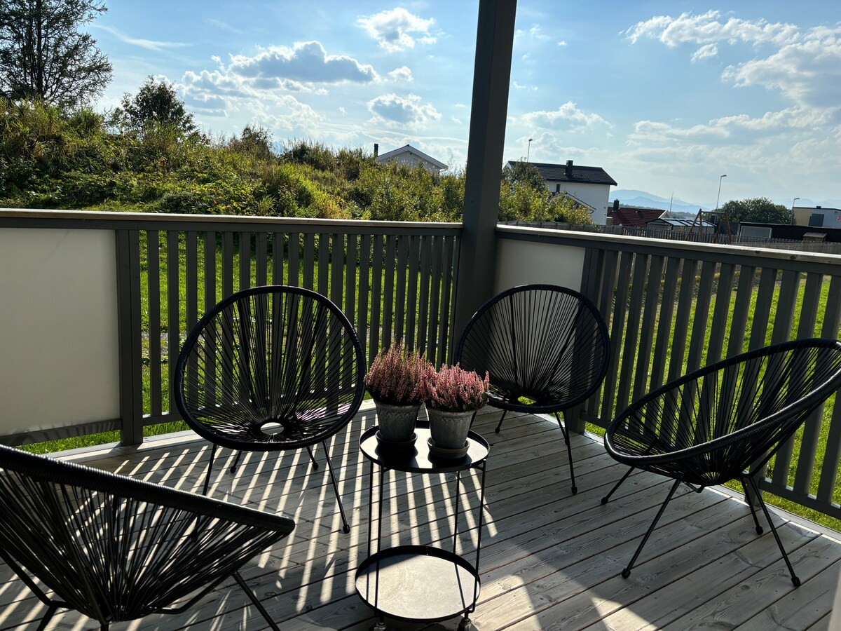 A spacious balcony features four black woven chairs arranged around a small table. Two potted plants with pink flowers are placed at the center. The surrounding view includes greenery and distant structures under a clear blue sky, highlighting a relaxing outdoor space.