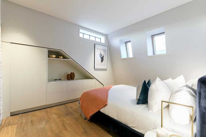 - Acurrúcate en una acogedora cama bajo un impresionante techo inclinado y rincones de decoración curados.
- Los tonos terrosos de la madera, las mantas de coral y las almohadas de felpa crean un ambiente ideal para relajarse.
- Deja que la suave luz de la mañana entre por las ventanas del trío para crear un ambiente relajante y elevado.