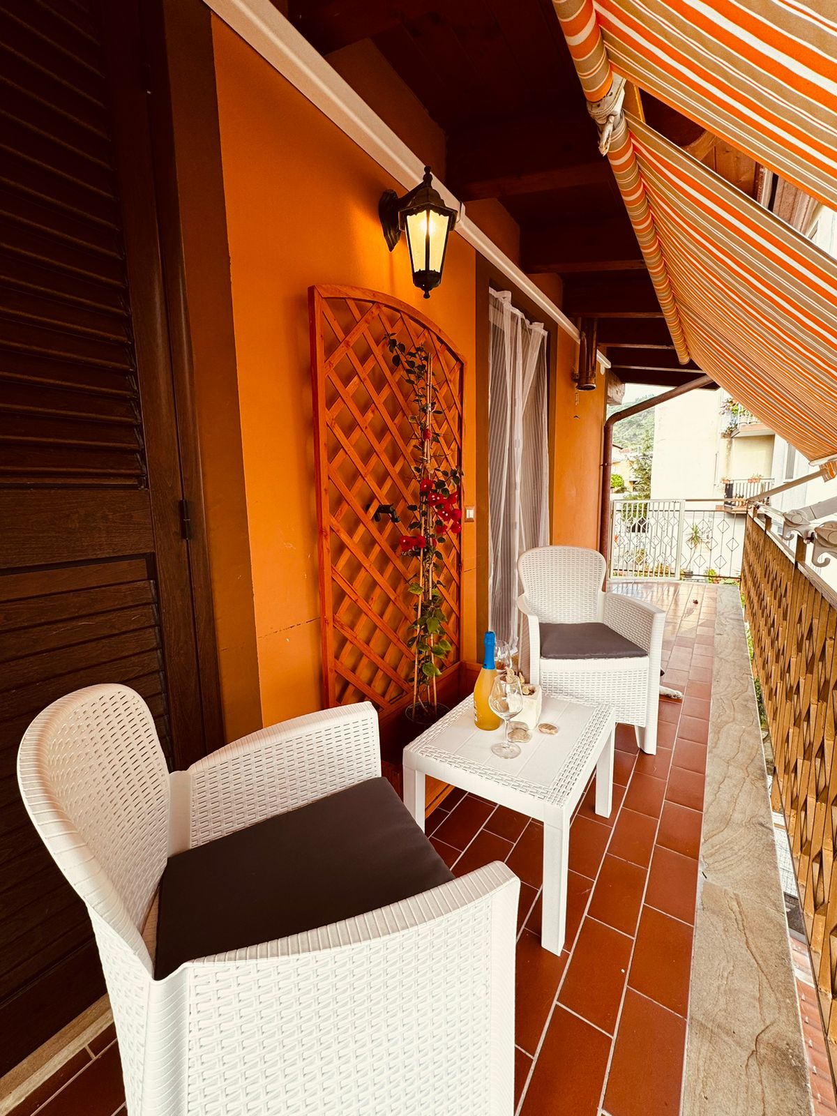 A cozy outdoor seating area features two white wicker chairs with dark cushions. A small table is positioned between them, set with two glasses and a bottle. Colorful striped awnings provide shade, while warm orange walls create a welcoming atmosphere.