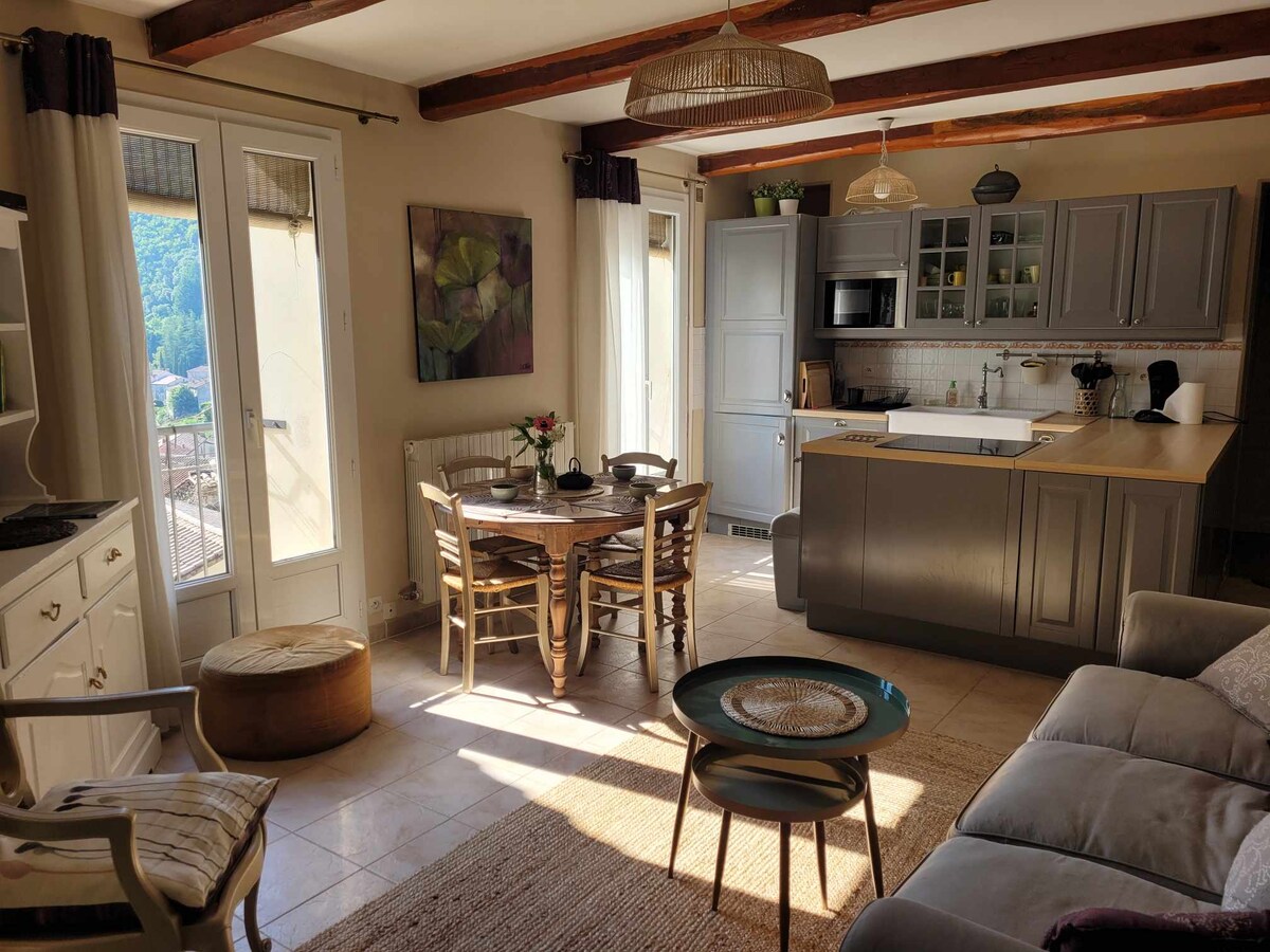 A combined living area features wooden beams overhead, enhancing the cozy atmosphere. A round dining table with four chairs is positioned near large windows, allowing natural light. The kitchen countertop, equipped with modern appliances, is visible, while a soft seating area includes a sofa and decorative elements.