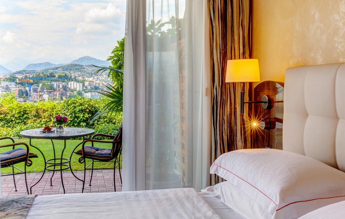 A bedroom is featured with a spacious bed positioned near a large window. A small outdoor terrace is visible, furnished with a round table and two chairs. Lush greenery and scenic views of the surrounding hills and city can be seen through the glass doors.