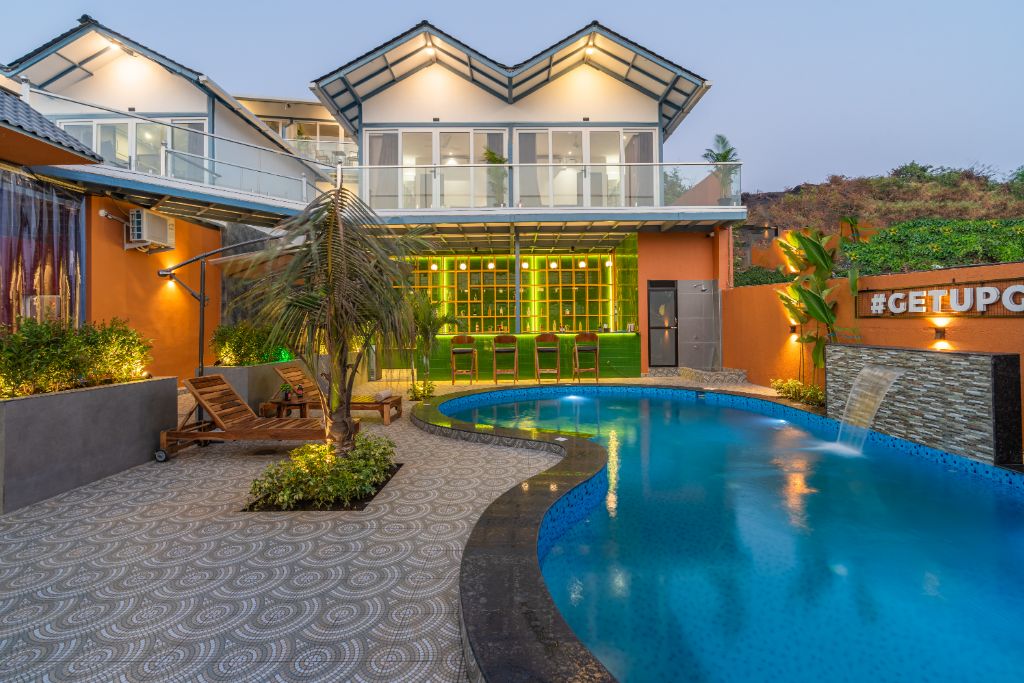 The outdoor area features a vibrant pool surrounded by lush greenery and lounge chairs. A bar with seating is visible in the background, under a modern roof design. The structure's large windows reflect the soft hues of twilight, creating a serene atmosphere.