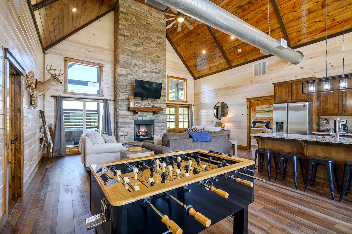 An open-concept living area features high ceilings with wooden beams, a stone fireplace, and large windows that provide natural light. A foosball table is centered in the space, surrounded by comfortable seating and a kitchen area with dark wood cabinetry and modern appliances.
