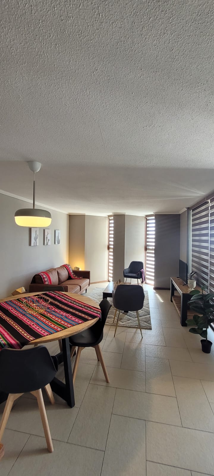 An open living area is highlighted by a colorful woven table runner across a dining table. Modern black chairs surround the table. A cozy seating area features a sofa, while a TV is positioned against a wall. Natural light filters through horizontal blinds.