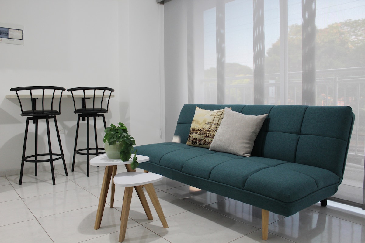 A modern living area features a teal sofa with a decorative pillow, accompanied by two small white tables. Behind, tall windows are adorned with sheer shades, allowing natural light to filter in. In the corner, two black bar stools are positioned at a small dining table.