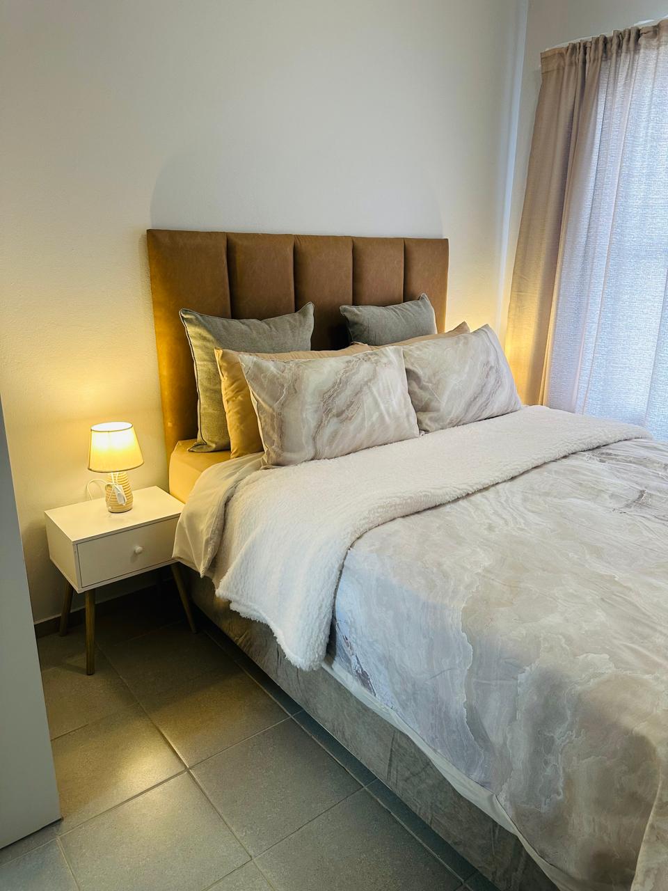 A cozy sleeping area features a spacious bed dressed in soft, neutral bedding and plush pillows. A bedside table holds a small lamp that casts a warm glow. Sheer curtains allow natural light to filter in, creating a serene atmosphere.