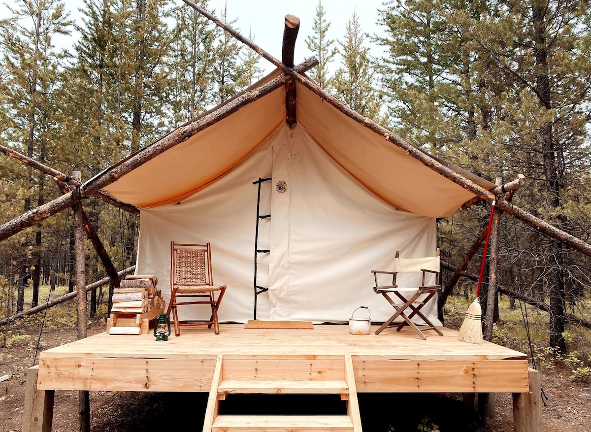 A cozy glamping tent is set atop a wooden deck, surrounded by trees. The entrance features a raised platform with two chairs and a small table, inviting guests to relax outdoors. A ladder leads to the tent interior, which offers rustic charm.
