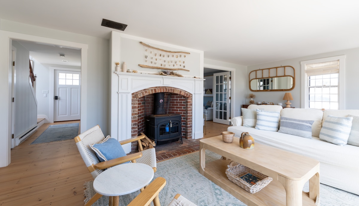 A light-filled living area features a comfortable sofa and wooden coffee table. A brick fireplace serves as a focal point, complemented by neutral decor. The space includes wooden accent chairs and a large mirror, enhancing the home's open and welcoming feel.