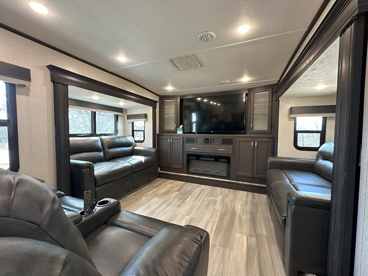 A spacious living area within the RV is showcased, featuring two comfortable leather sofas positioned across from a large television. Natural light filters in through several windows, illuminating the neutral-toned walls and sleek flooring.
