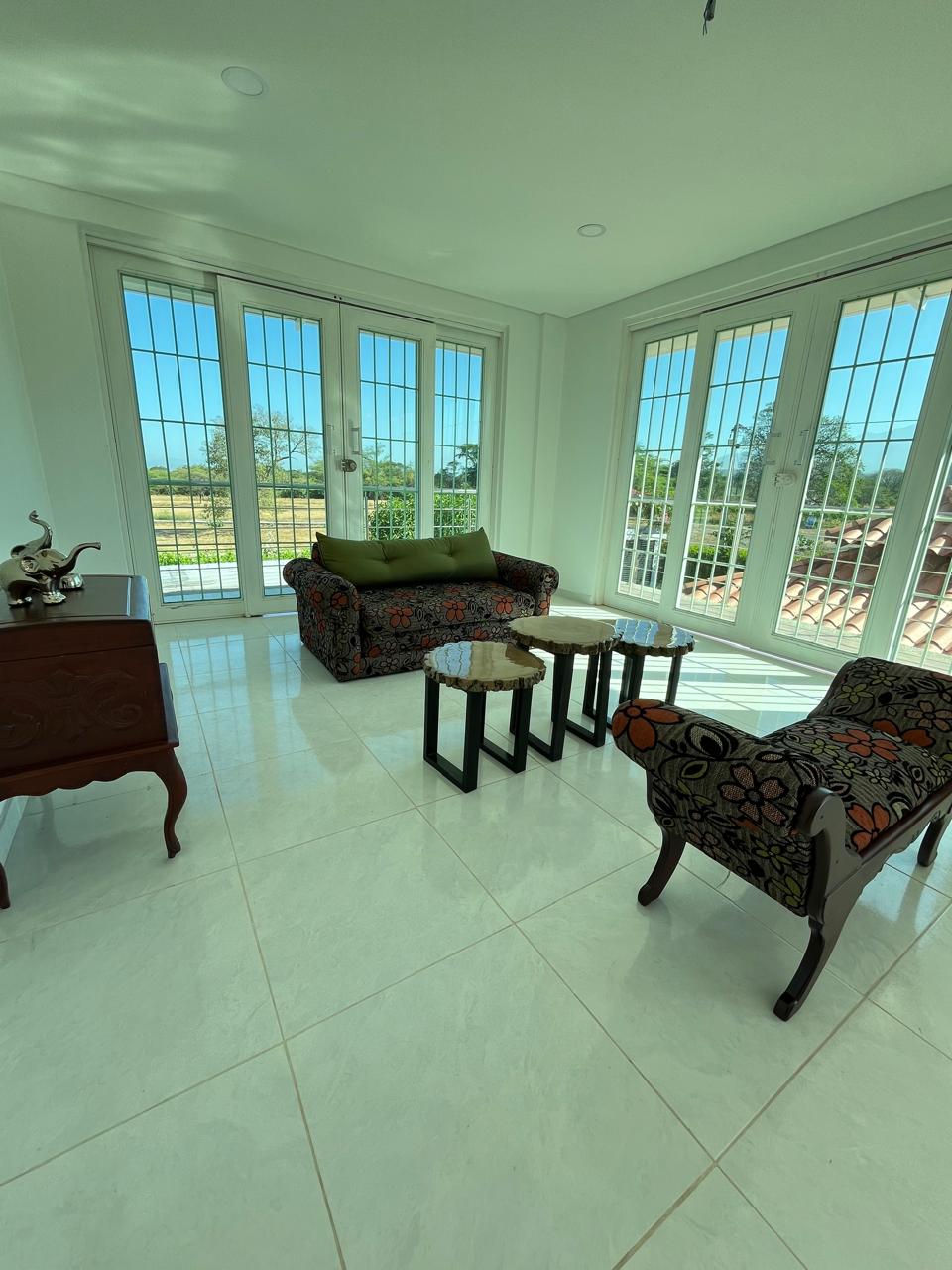 A bright seating area features large windows that allow ample natural light. A patterned sofa and two small side tables are arranged on a glossy tiled floor, with a decorative cabinet visible in the corner. The outdoor greenery is visible through the windows.