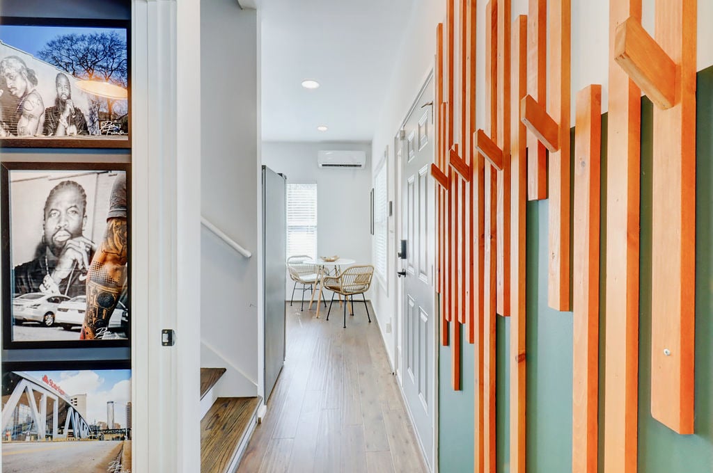 A well-lit hallway features wooden wall art that adds a creative touch. Framed photographs decorate one wall, showcasing local scenes. At the end of the hallway, a dining area is visible, containing a small round table and four chairs, inviting casual meals and gatherings.