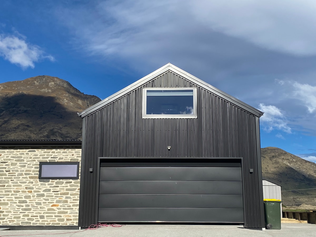 The exterior of a modern building features a sleek black corrugated facade with a prominent dark garage door. A large window is situated centrally near the peak of the roof, offering natural light. The surrounding landscape includes hills under a cloudy sky.